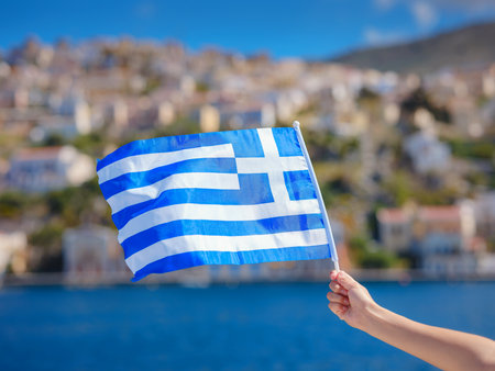 View of Greek flag waving in air serene Mediterranean vibe. colorful neoclassical houses in harbor town of Symi (Symi Island, Greece)の写真素材