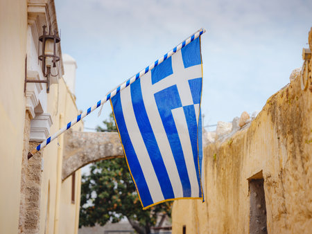View of flag waving in air serene Mediterranean vibe. beautiful Greek islands series, Narrow cobblestone street lined with charm in downtown Rhodes , Greece. Idyllic stone alleyの写真素材
