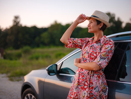Traveler woman in red dress and hat drives car through scenic countryside of Croatia in summer. Surrounded by lush fields and forests, she enjoys peaceful and adventurous road tripの写真素材