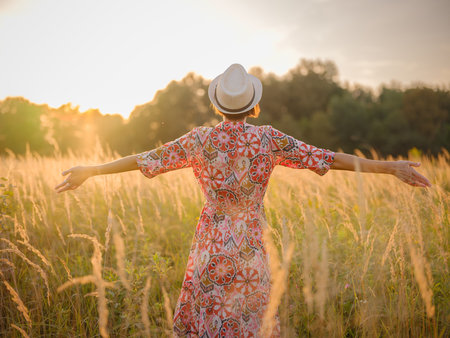 Young woman walking through picturesque European field in late summer. Golden sunlight, lush greenery, and serene rural atmosphere create a peaceful countryside scene.の写真素材