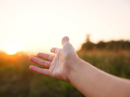 Soft evening light bathes golden field as woman's hand gently moves through tall grass, capturing the sense of freedom, nature, and serenity. Warm sunset hues create a peaceful, dreamy countryside atmosphere.の写真素材