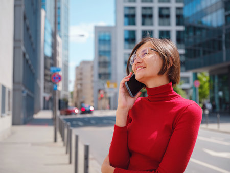 Woman in glasses and red turtleneck, speaks on phone in center of Frankfurt, surrounded by modern buildings and fast-paced city life, balancing professionalism and communication in urban setting.の写真素材