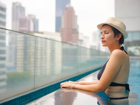 Young Asian woman relaxing by pool at Kuala Lumpur hotel with view of surrounding skyscrapers, enjoying leisure time in vibrant urban setting.の写真素材