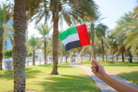 Close up of female hand holding tiny flag of UAE against Abu Dhabi park from Corniche. Promenade in Abu Dhabi. United Arab Emirates, Middle East. Photo of UAE national day celebration spirit.の写真素材