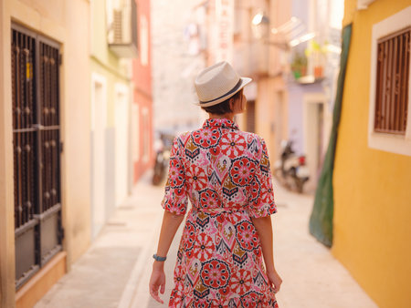 Woman in dress strolls through colorful streets of Spanish coastal town of La Vila Joiosa. sunny winter highlights atmosphere charm of Mediterranean architecture and quiet seaside life, back viewの写真素材