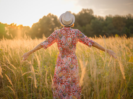 Young woman walking through picturesque European field in late summer. Golden sunlight, lush greenery, and serene rural atmosphere create a peaceful countryside scene.の写真素材