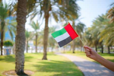 Close up of female hand holding tiny flag of UAE against Abu Dhabi park from Corniche. Promenade in Abu Dhabi. United Arab Emirates, Middle East. Photo of UAE national day celebration spirit.の写真素材
