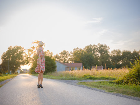 Young woman walking through picturesque European field in late summer. Golden sunlight, lush greenery, and serene rural atmosphere create a peaceful countryside scene.の写真素材
