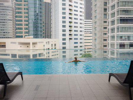Young asian woman relaxing by pool at Kuala Lumpur hotel with view of surrounding skyscrapers, enjoying leisure time in vibrant urban setting.の写真素材