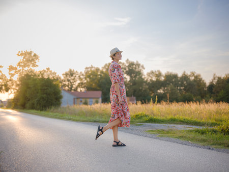 Young woman walking through picturesque field in late summer. Golden sunlight, lush greenery, and serene rural atmosphere create peaceful countryside scene.の写真素材
