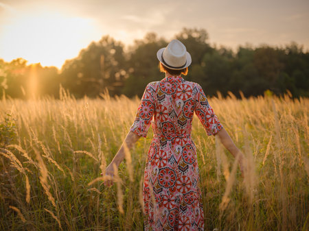 Young woman walking through picturesque European field in late summer. Golden sunlight, lush greenery, and serene rural atmosphere create peaceful countryside scene.の写真素材