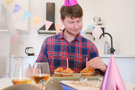 man celebrates with four cupcakes topped with candles in place of traditional birthday cake. Smiling and joyful, he enjoys cozy and unique celebration, creating warm and memorable festive moment.の写真素材