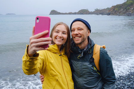 Couple enjoys taking selfie on beach in Winter. walking on stones, watching sea waves, relaxing together, slow romantic moment near calm blue water under soft winter light.の写真素材
