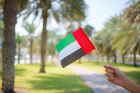 Close up of female hand holding tiny flag of UAE against Abu Dhabi park from Corniche. Promenade in Abu Dhabi. United Arab Emirates, Middle East. Photo of UAE national day celebration spirit.の写真素材