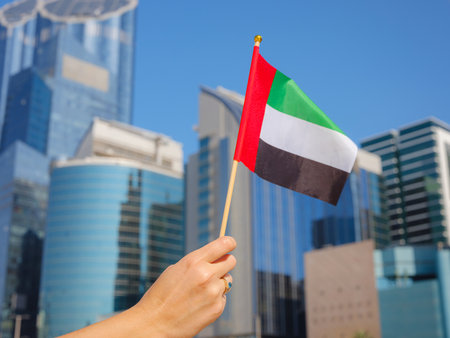 Close up of female hand holding tiny flag of UAE against Abu Dhabi skyline. Photo of UAE national day celebration spirit.の写真素材