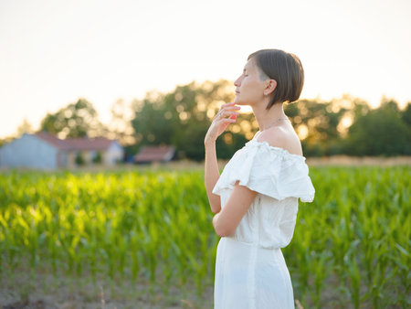 Beautiful woman in white dress standing in summer field during golden sunset, warm sunlight, romantic evening mood, natural countryside atmosphere, soft wind and peaceful sceneの写真素材
