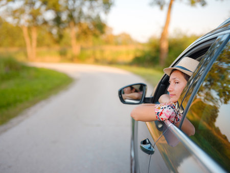 Traveler woman in red ethnic dress and hat drives car through scenic countryside of Croatia in summer. Surrounded by lush fields and forests, she enjoys peaceful and adventurous road tripの写真素材