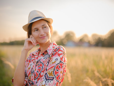 Young woman walking through picturesque European field in late summer. Golden sunlight, lush greenery, and serene rural atmosphere create peaceful countryside scene.の写真素材