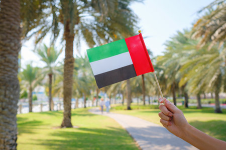 Close up of female hand holding tiny flag of UAE against Abu Dhabi park from Corniche. Promenade in Abu Dhabi. United Arab Emirates, Middle East. Photo of UAE national day celebration spirit.の写真素材