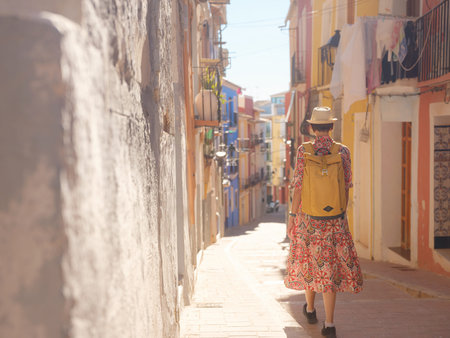Woman in dress strolls through colorful streets of Spanish coastal town of La Vila Joiosa . sunny winter atmosphere highlights charm of Mediterranean architecture and quiet seaside life, back viewの写真素材