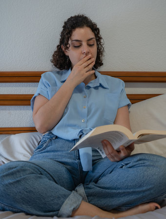 Woman yawns while reading a book sitting on bedの写真素材