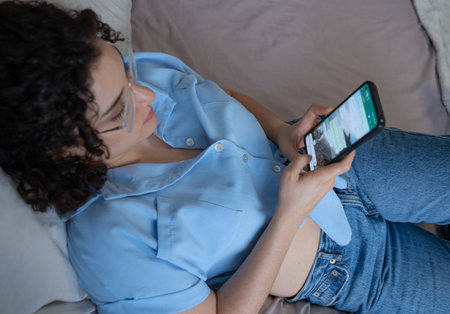 Woman texts on mobile while lying on bedの写真素材