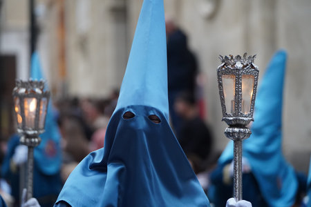 Three Nazarenes in blue, two of them carrying a candle lit with a candle, on the city street while...