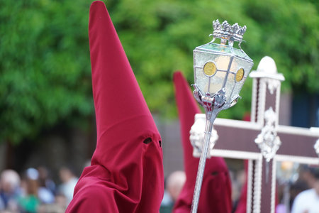 A red-colored Nazarene, in a row, with a lantern and a candle inside, and the guiding cross at its side, processes through the streets of the city during Holy Week.の写真素材
