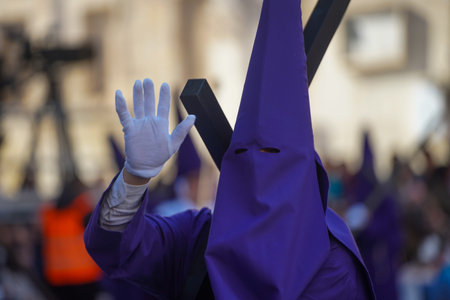 Nazarene in purple, with white gloves and carrying a cross, greets the camera that photographs him,...