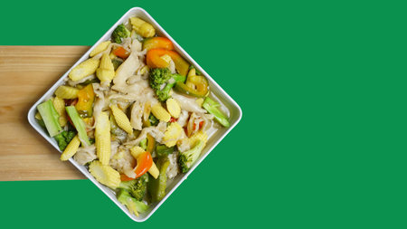 Young corn capcay vegetables with mushrooms and also peppers are seen from the above on a white box plate against a green backgroundの写真素材