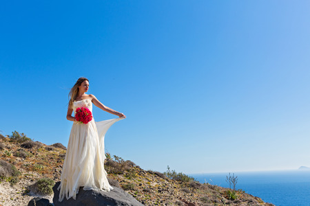 Beautiful woman in white dress stands on a high stone near the seaの写真素材