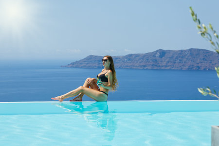 Young woman sitting on the edge of the pool, back to the sea and drinking a cocktail