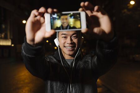 Young asian man with headphones taking a selfie with smart phone in the city.の写真素材