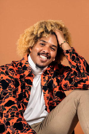 Stock photo of cheerful afro man smiling and looking at camera in studio shot against brown...
