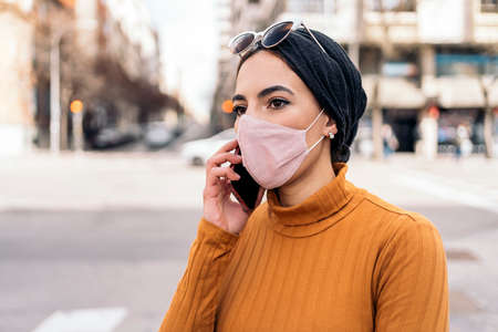 Expressive young muslim woman wearing protective face mask using her mobile phone in the street.の写真素材
