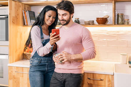 Beautiful black woman showing her boyfriend something in the phone while standing in the kitchen at home.の写真素材