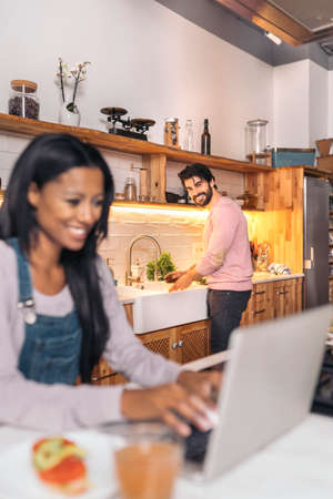 Beautiful black woman using her laptop in the kitchen while her boyfriend cooks.の写真素材