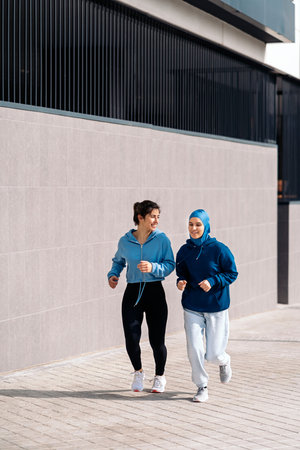 Active young female friends running in the street and smiling. One of them is wearing a hijab.