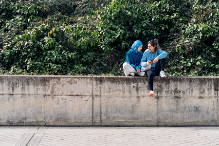Cheerful muslim woman wearing hijab and sports clothes sitting in the street with her friend and using mobile phone.の写真素材