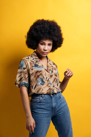 Serious afro woman wearing colorful shirt posing and looking at camera in studio shot against yellow background.の写真素材