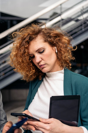 Concentrated business woman with curly hair and beautiful suit using her mobile phone and listening to music.の写真素材
