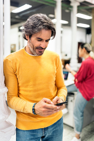 Smiley male office worker using his mobile phone and standing in the office. Coworking concept.