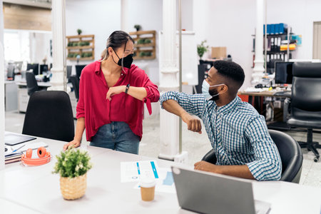 Young asian office worker talking with her african colleague and doing elbow bump. They are wearing face masks due to covid19.の写真素材