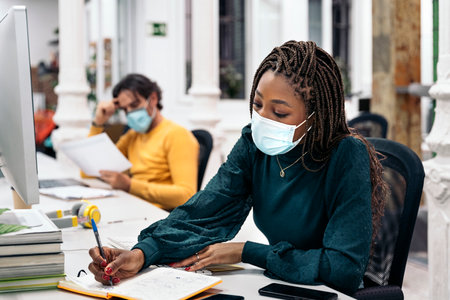 Focused black woman with face mask sitting in her desk and doing paper work next to other coworkers. Office concept.の写真素材