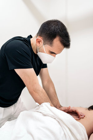 Professional worker in physiotherapy clinic wearing face mask working with unrecognized client.