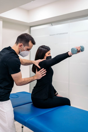 Male physiotherapist wearing face mask working with young woman in physiotherapy clinic.