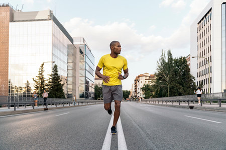 Front view of an African American athlete man with yellow t-shirt and shorts jogging on the middle of the street in the city outdoors.の写真素材