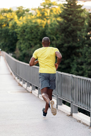 Rear view of an african american sportsman with yellow t-shirt and shorts jogging on sidewalk in the...