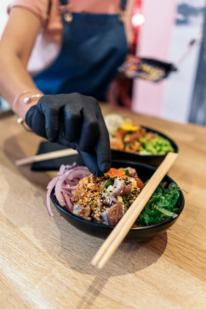 Unrecognized restaurant worker in protective gloves putting ingredients into poke bowl with rice and...