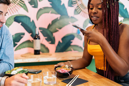 Pretty african woman with braids enjoying poke bowl with her friend and taking a photo of her food.